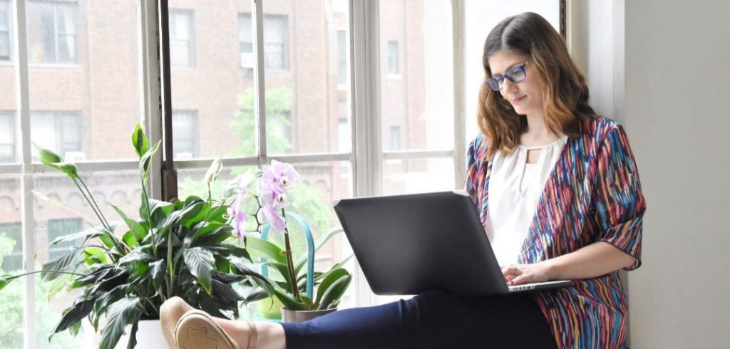 Freelance writer sitting on window ledge using laptop to make website